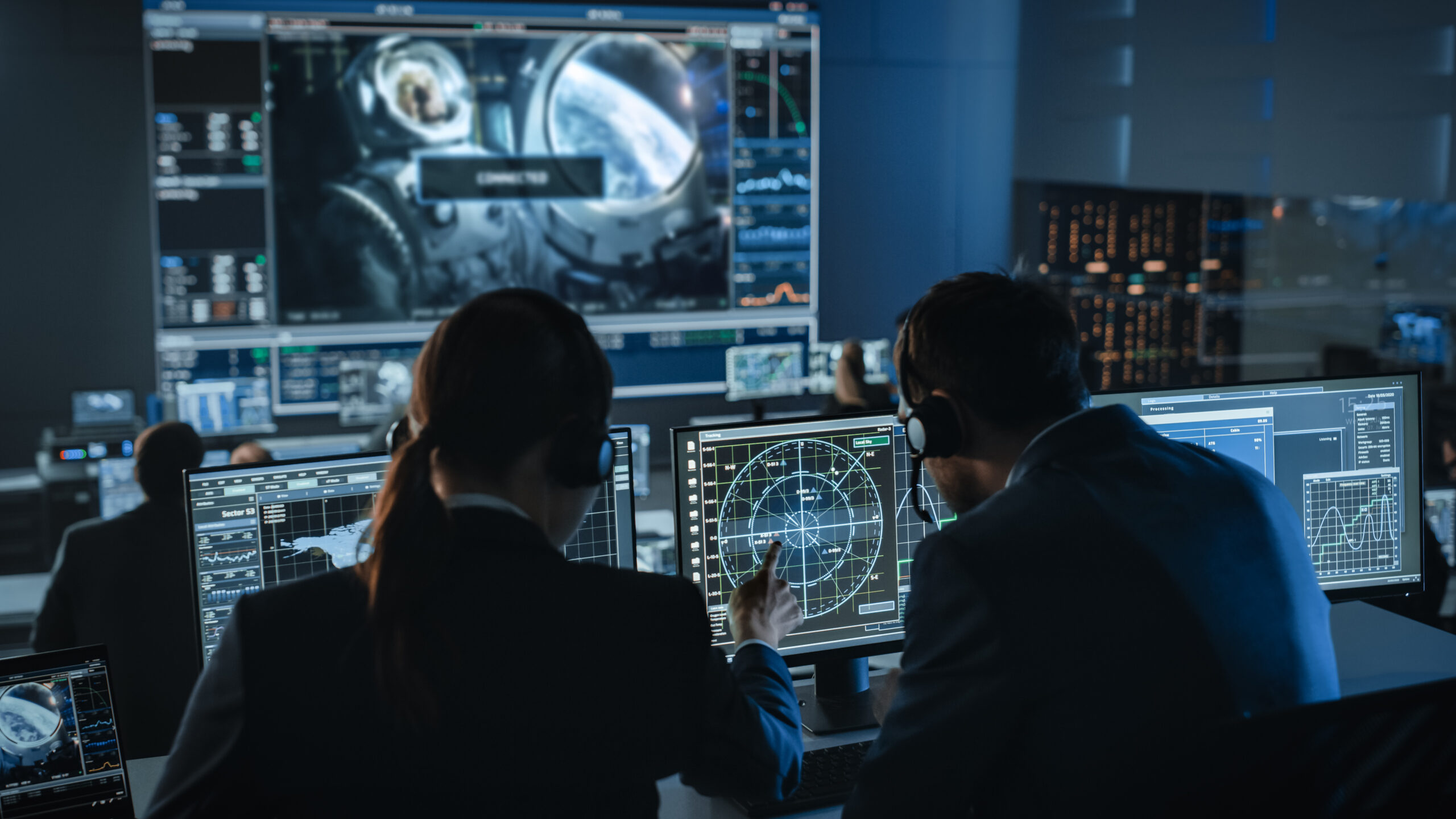 Group of People in Mission Control Center Establish Successful Video Connection on a Big Screen with an Astronaut on Board of a Space Station. Flight Control Scientists Sit in Front Computer Displays.